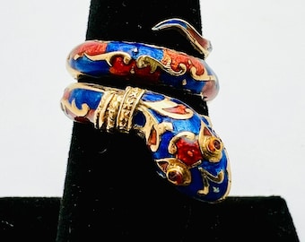 May include: A gold-toned snake ring with blue, red, and gold enamel detailing. The ring is coiled, with the snake's head and tail overlapping. The design features intricate patterns and a polished finish, showcasing a unique jewelry piece.