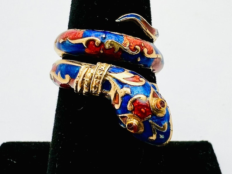 May include: A gold-toned snake ring with blue, red, and gold enamel detailing. The ring is coiled, with the snake's head and tail overlapping. The design features intricate patterns and a polished finish, showcasing a unique jewelry piece.