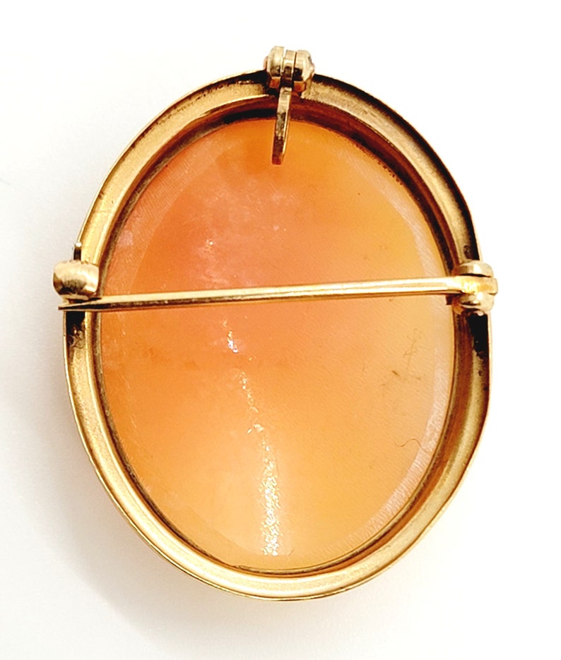 May include: An oval cameo brooch in a gold tone with a light orange cameo stone. The brooch has a pin and catch fastening on the reverse.