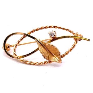 Op de afbeelding: Een gouden broche met een bladontwerp en een enkele diamant accent. Het blad is getextureerd en de broche is gemaakt van gedraaid draad.