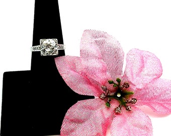 Könnte beinhalten: Ein silberner Ring mit einem großen, runden, klaren Edelstein, der von kleineren, klaren Edelsteinen umgeben ist. Der Ring befindet sich auf einem schwarzen Ringständer mit einer rosa Blume.