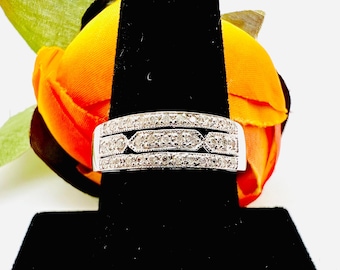 May include: A silver ring with three rows of small, clear gemstones. The center row features an alternating geometric design. The ring is displayed on a black velvet surface, with a colorful orange and yellow flower in the background.