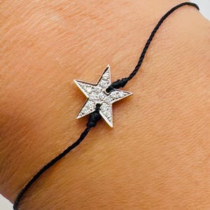 Könnte beinhalten: Ein Armband mit einem silbernen Sternanhänger und einem schwarzen Band. Der Stern ist mit kleinen, klaren Kristallen besetzt. Das Armband wird am Handgelenk einer Person gezeigt.
