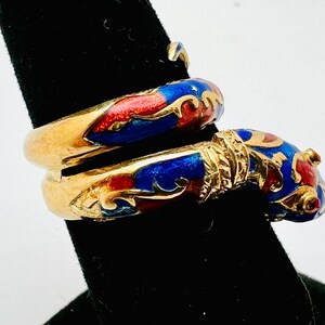 May include: A gold-toned ring with a wrap-around design. The ring features red and blue enamel detailing. The enamel is accented with gold-colored filigree. The ring is displayed on a black surface.
