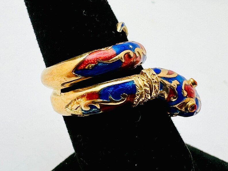 May include: A gold-toned ring with a wrap-around design. The ring features red and blue enamel detailing. The enamel is accented with gold-colored filigree. The ring is displayed on a black surface.