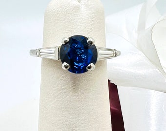 May include: A silver ring with a large, oval-shaped blue sapphire gemstone surrounded by two smaller, baguette-shaped diamonds.