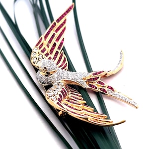 Puede incluir: Un broche de tono dorado en forma de golondrina, con piedras rojas y transparentes. Las alas de la golondrina están extendidas y su cola está extendida.