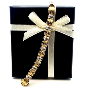 May include: A gold and white diamond bracelet with a textured design. The bracelet is displayed in a black gift box with a white ribbon.