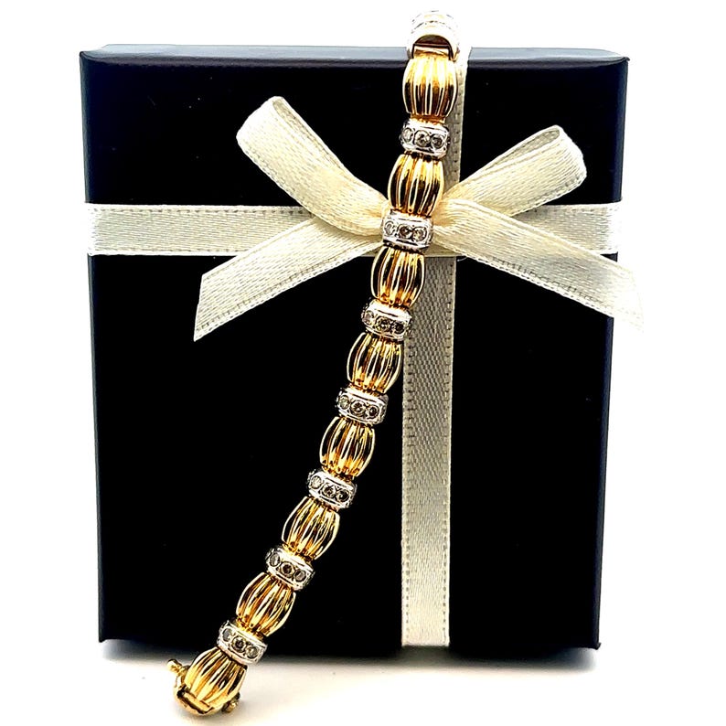 May include: A gold and white diamond bracelet with a textured design. The bracelet is displayed in a black gift box with a white ribbon.