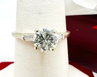 Puede incluir: Un anillo de plata con un gran diamante redondo y claro en el centro y dos diamantes más pequeños en forma de baguette a cada lado.
