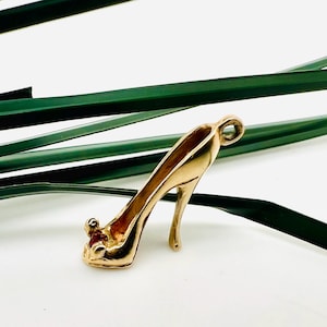 May include: A gold-colored high-heeled shoe charm with a loop for a necklace or bracelet. The charm is set against a white background with green leaves.