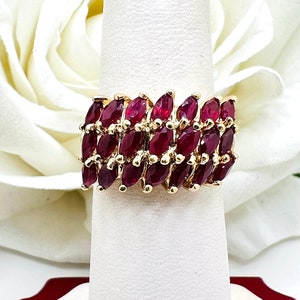 Könnte beinhalten: Ein goldener Ring mit einer Reihe von rubinroten Steinen in Marquise-Form. Der Ring befindet sich auf einem weißen Präsentationsständer.