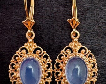 May include: A pair of gold-colored drop earrings. Each earring features an oval, light blue stone set within an ornate, filigree-style frame. The earrings have a lever-back closure.