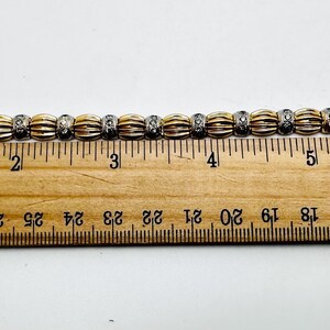 May include: A gold and silver bracelet with a row of round, diamond-like stones. The bracelet is measured on a ruler, showing it is approximately 6 inches long.
