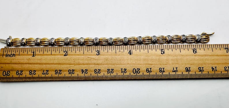 May include: A gold and silver bracelet with a row of round, diamond-like stones. The bracelet is measured on a ruler, showing it is approximately 6 inches long.