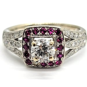 Pode incluir: Um anel de noivado de prata com um grande diamante redondo central rodeado por um halo de rubis. A banda é decorada com diamantes menores.