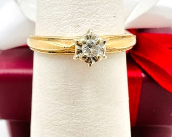 Könnte beinhalten: Ein Goldring mit einem einzelnen runden Diamanten in einer Krappenfassung. Der Ring befindet sich auf einem weißen Präsentationsständer.