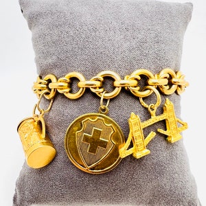 May include: A gold charm bracelet with a chain link design and multiple charms, including a Swiss shield, a beer stein, and a miniature replica of the Tower Bridge.