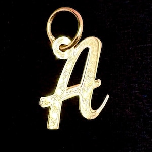 May include: A gold-colored pendant in the shape of the letter "A" with a loop at the top for a chain. The pendant has a textured surface and is set against a black background. This jewelry item is suitable for necklaces and other accessories.