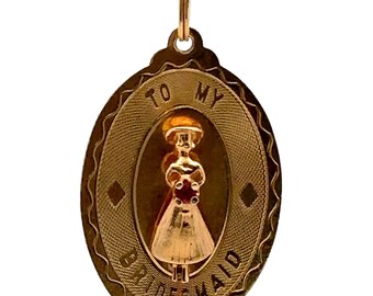 Puede incluir: Collar con colgante dorado con un dije ovalado. El dije presenta un diseño en relieve con las palabras "TO MY BRIDESMAID" y una figura central de una mujer con un vestido. La cadena del collar también es dorada.