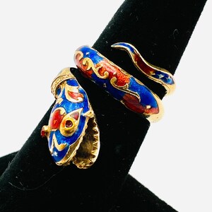 May include: A gold-toned snake ring with blue, red, and white enamel detailing. The ring is wrapped around a black display stand, showcasing the intricate design of the serpent's head and body. The ring's design is reminiscent of vintage jewelry.
