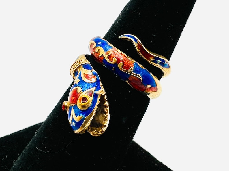 May include: A gold-toned snake ring with blue, red, and white enamel detailing. The ring is wrapped around a black display stand, showcasing the intricate design of the serpent's head and body. The ring's design is reminiscent of vintage jewelry.