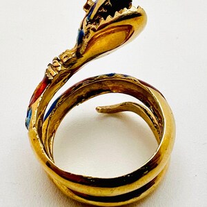 May include: A gold-colored snake ring with red, blue, and black enamel detailing. The snake's head is raised, with its mouth open, and the body coils around the finger. The ring is set against a white background.
