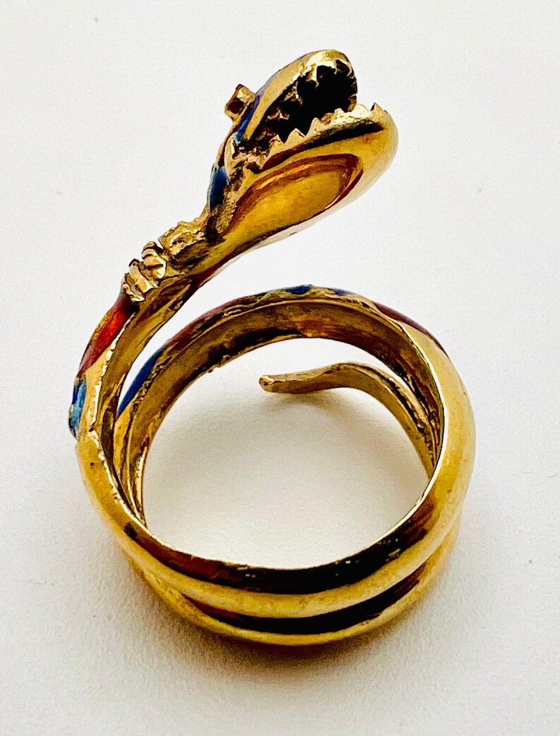 May include: A gold-colored snake ring with red, blue, and black enamel detailing. The snake's head is raised, with its mouth open, and the body coils around the finger. The ring is set against a white background.