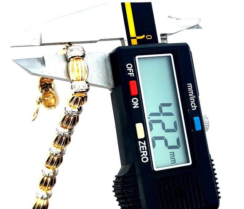 May include: A close-up of a gold and white diamond bracelet being measured with a digital caliper. The caliper reads 12.2 mm.