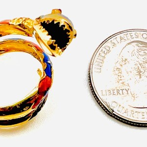 May include: Gold-toned ring with a shark design, featuring black, red, and blue enamel accents. The ring is next to a US quarter for size comparison. The shark's mouth is open, revealing black teeth.