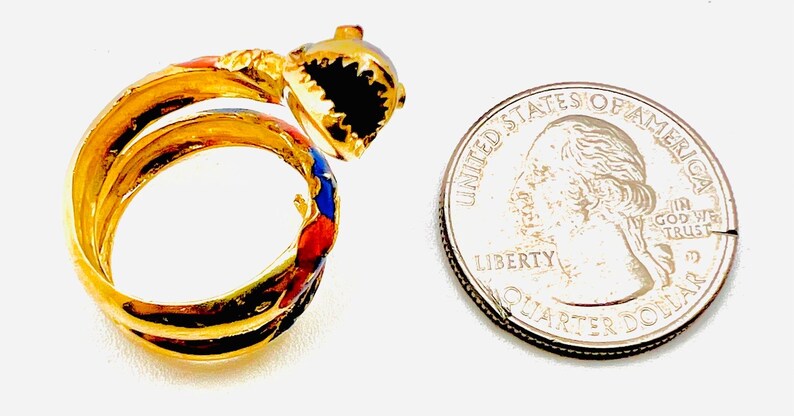 May include: Gold-toned ring with a shark design, featuring black, red, and blue enamel accents. The ring is next to a US quarter for size comparison. The shark's mouth is open, revealing black teeth.