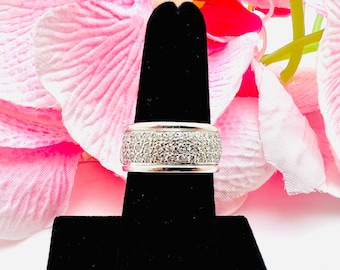 May include: A wide, silver-toned ring with a band encrusted with small, clear gemstones. The ring is displayed on a black stand against a backdrop of pink and red flowers. The ring's design is simple yet elegant.