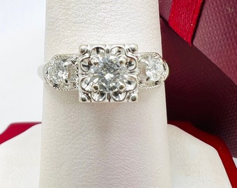 Puede incluir: Un anillo de compromiso de plata con un diamante central de forma cuadrada y dos diamantes más pequeños a cada lado.