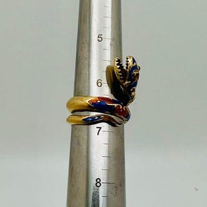 May include: A gold-toned snake ring with red, blue, and white enamel detailing. The snake's head is raised, and the ring is wrapped around a metal sizing tool. The ring's design is intricate and detailed.