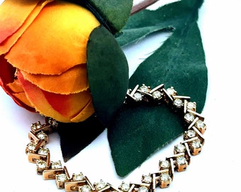 May include: A delicate gold bracelet with a chevron pattern set with sparkling clear stones. The bracelet is lying on a green leaf and a yellow flower.