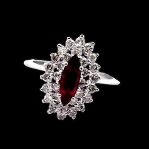 May include: A silver ring featuring a marquise-cut, deep red gemstone surrounded by a halo of small, round, clear gemstones. The ring's band is thin and polished, set against a black background.