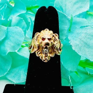 Könnte beinhalten: Ein goldfarbener Ring mit einem Löwenkopf-Design. Der Löwe hat rote Augen und einen kleinen Diamanten im Maul. Der Ring wird auf einem schwarzen Ständer vor einem türkisfarbenen Stoffhintergrund präsentiert.