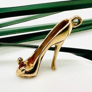 May include: A gold-colored high heel charm with a small red accent on the toe. The charm is detailed with a curved heel and a loop at the top for attaching to a necklace or bracelet. The background includes green leaves.
