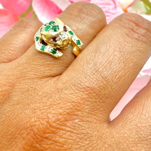 May include: A gold ring with a panther design, set with emerald and ruby gemstones. The panther's eyes are ruby, and the body is accented with emeralds and small clear stones. The ring is worn on a finger, with a blurred floral background.