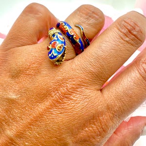 May include: Two ornate rings with a snake design. One ring is blue with red and gold accents, and the other is red and blue with gold accents. The rings are on a hand, showcasing their intricate details and vibrant colors.