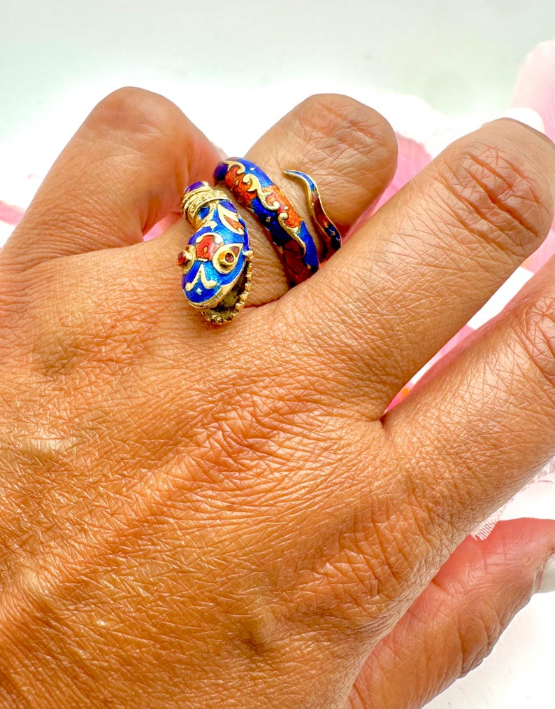 May include: Two ornate rings with a snake design. One ring is blue with red and gold accents, and the other is red and blue with gold accents. The rings are on a hand, showcasing their intricate details and vibrant colors.