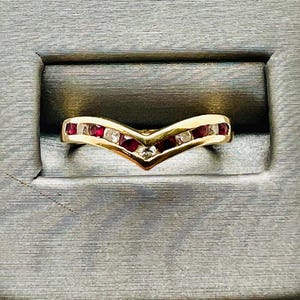 May include: A gold-toned ring with a V-shaped design. The ring is set with alternating red and clear gemstones. The gemstones are small and evenly spaced along the band. The ring is displayed on a gray surface.