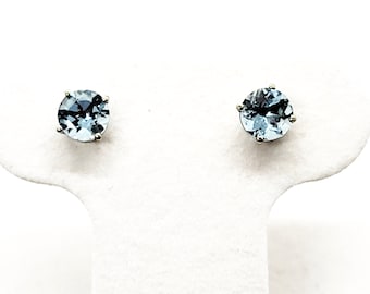 May include: A pair of stud earrings featuring round, light blue gemstones set in a silver-colored metal. The earrings are displayed on a white surface, showcasing their color and design. The gemstones have a faceted appearance.