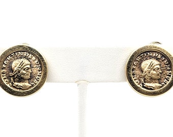 May include: A pair of gold-toned earrings featuring a Roman coin design. Each earring features a profile of a person wearing a helmet with the inscription "IMP CAESAR AVG F AVGVSTVS" around the edge.