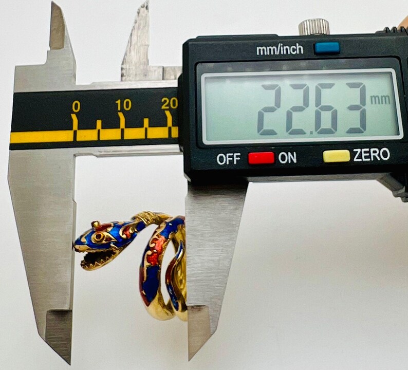 May include: A gold-colored snake ring with blue and red enamel detailing is measured with a digital caliper. The ring's head is open-mouthed, and the caliper displays a measurement of 22.63 mm.