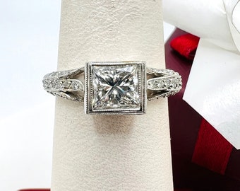 May include: A square-cut diamond engagement ring set in a platinum band with a delicate, intricate design. The ring is displayed on a white ring stand.