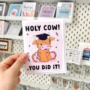 May include: A pink and white greeting card with a cartoon illustration of a brown highland cow wearing a graduation cap and holding a diploma. The text on the card reads "HOLY COW! YOU DID IT!"