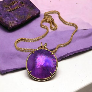 Könnte beinhalten: Eine goldfarbene Halskette mit einem großen, runden, facettierten lila Anhänger. Der Anhänger ist in einen goldenen Rahmen gefasst und hängt an einer zarten Goldkette. Ein lila quadratisches Objekt befindet sich im Hintergrund.