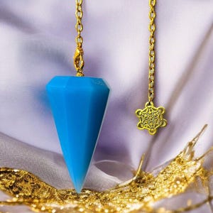 Könnte beinhalten: Ein facettiertes, hellblaues Pendel mit einer goldenen Kette und einem Verschluss. Eine zweite goldene Kette hält einen dekorativen, durchbrochenen Anhänger. Das Pendel liegt auf einem weichen, drapierten Stoff mit goldenen Akzenten.