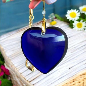Puede incluir: Un collar con colgante en forma de corazón con un centro azul oscuro y un borde negro. El colgante cuelga de una delicada cadena dorada. El collar está sostenido por una mano con uñas cuidadas. Las margaritas y las flores rosas están en el fondo.